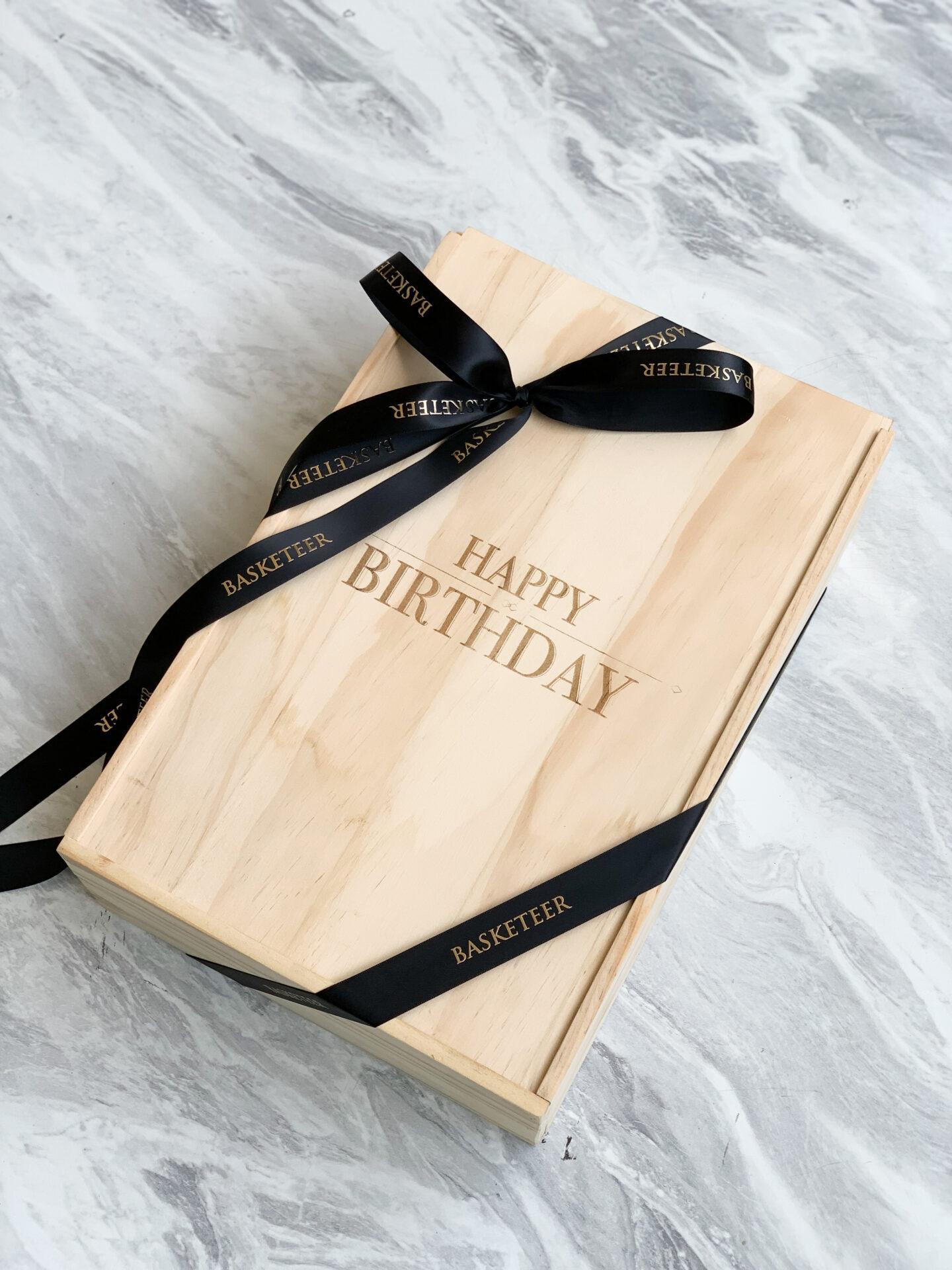 A light wooden gift box sits on a grey marble surface. The box has "Happy Birthday" engraved on the top and is wrapped with a black ribbon that reads "Basketeer." The ribbon is tied in a bow at the center of the box.