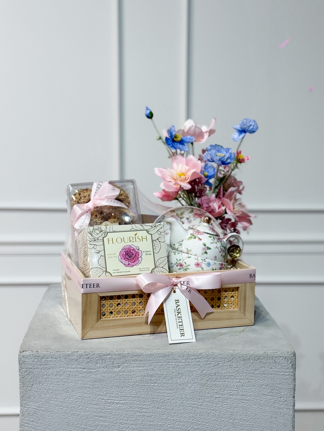 A wooden gift box with a pink ribbon holds a floral teacup set, biscuits, and tea—The Perfect Mother’s Day Pampering. A bouquet of pink and blue flowers in a teapot sits beside the box, all displayed on a grey pedestal.