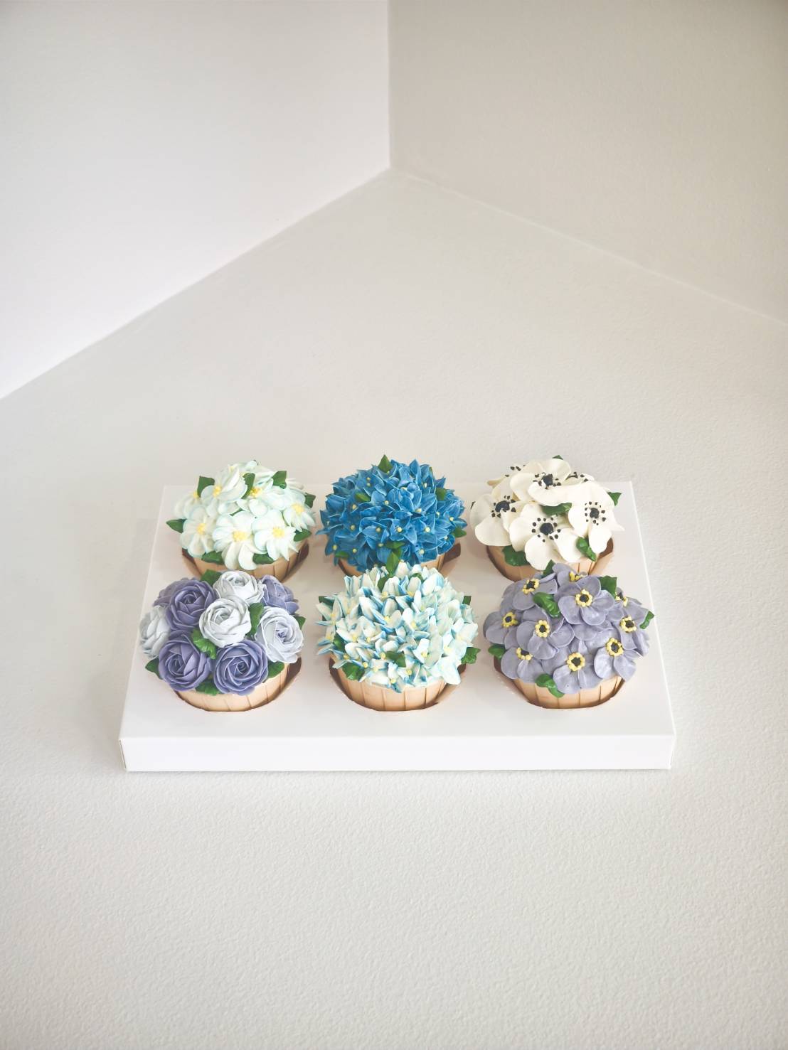 Elegant set of six floral cupcakes decorated with intricate buttercream flowers in shades of blue, purple, and white.