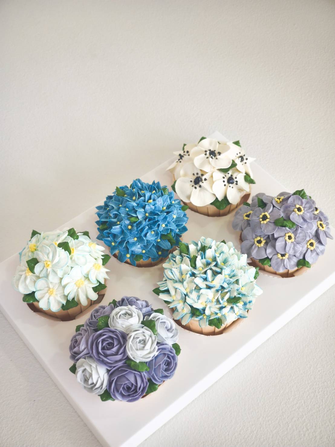 Set of six luxury cupcakes decorated with hand-piped buttercream flowers in shades of blue, white, and purple.