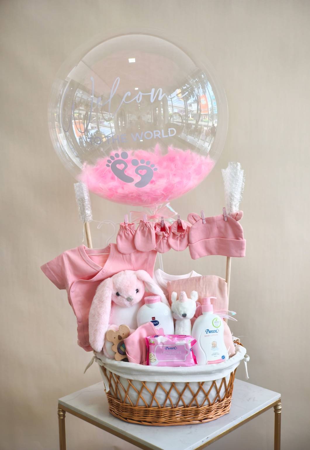 Luxury pink baby gift basket with baby clothes, plush toys, Pureen baby care products, and balloon with ‘Welcome to the World’ message.