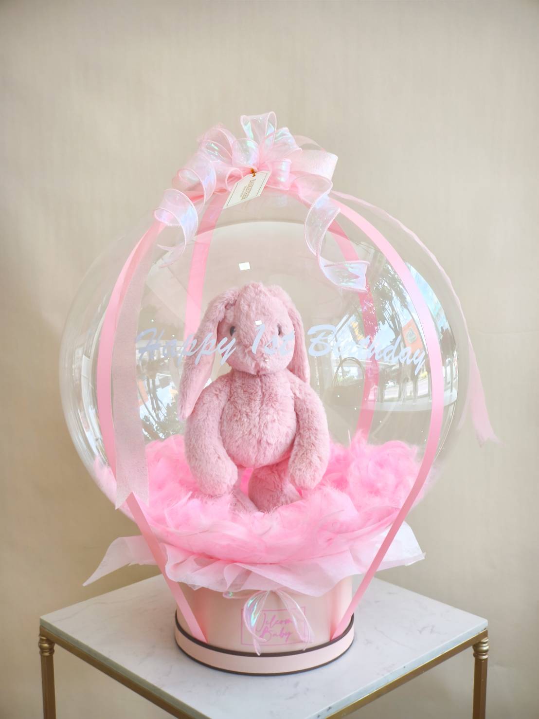 Clear balloon surprise with pink bunny plush toy, pink ribbon, and fluffy base in pastel pink box.