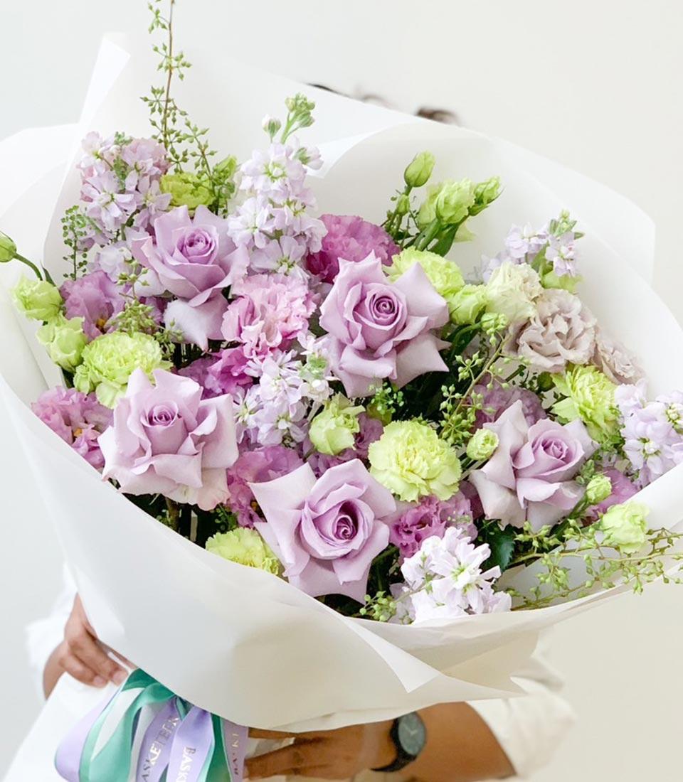 A large, elegant bouquet with a mix of light purple roses, pink flowers, and green carnations surrounded by smaller white flowers and greenery, wrapped in white paper. The bouquet is being held by a person with only their hands visible.