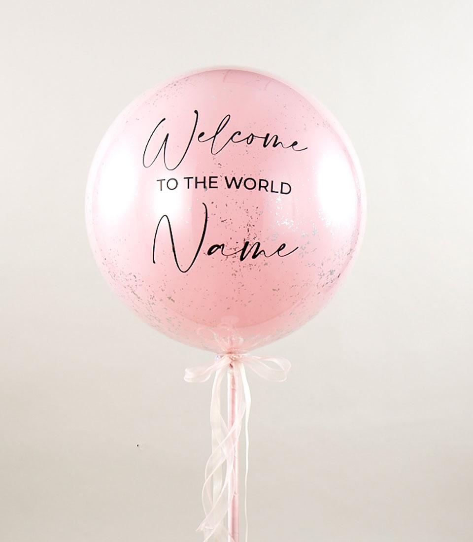 A large, round, pink balloon with a light sprinkling of sparkles and a white ribbon tied at the bottom. This personalized pink welcome balloon gift features the text 