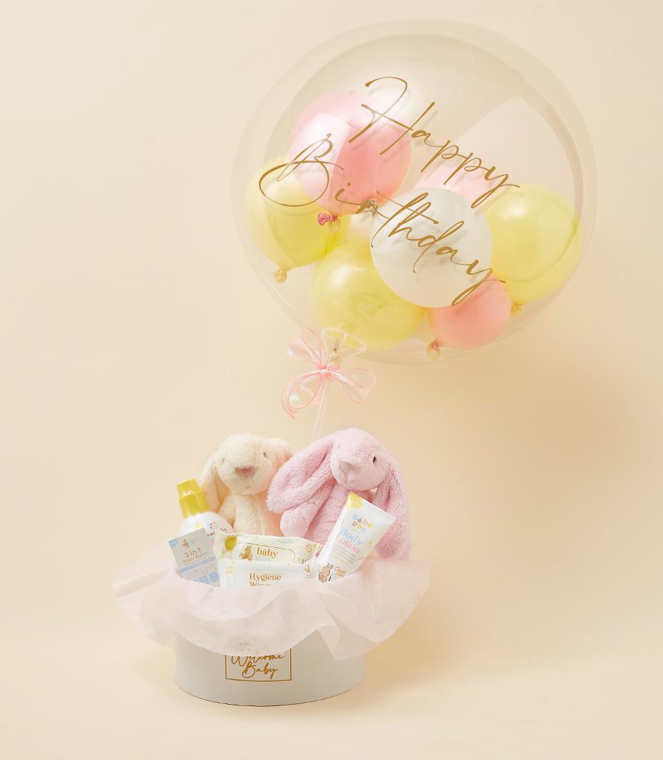 A round gift box contains a variety of baby products including two plush bunnies, baby lotion, a baby bottle, and diapers. Above the box, a clear balloon filled with pink and yellow balloons bears the text 