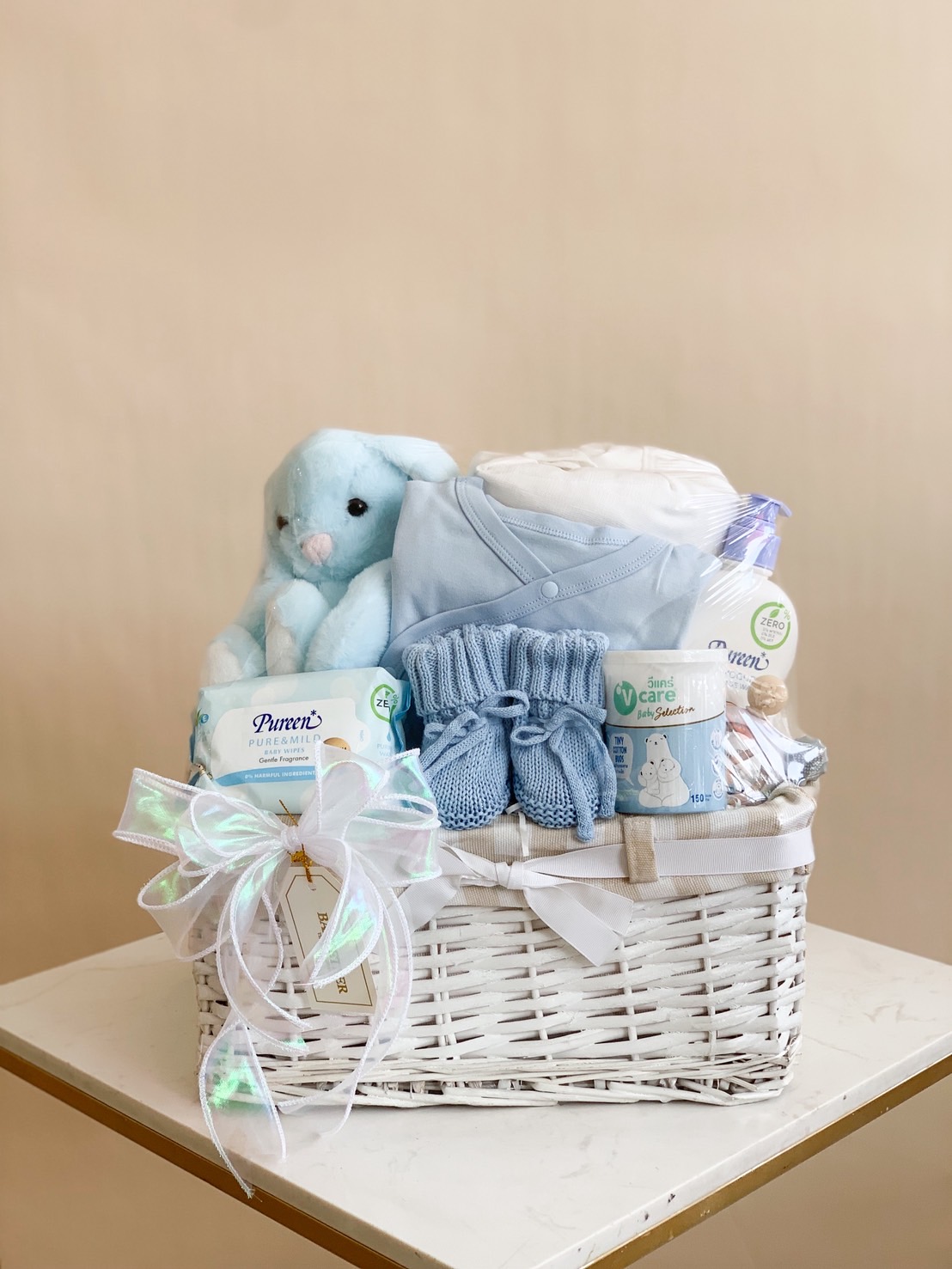 Blue baby boy gift basket with blue bunny plush, baby clothes, booties, wipes, and lotion in a white wicker basket with ribbon.
