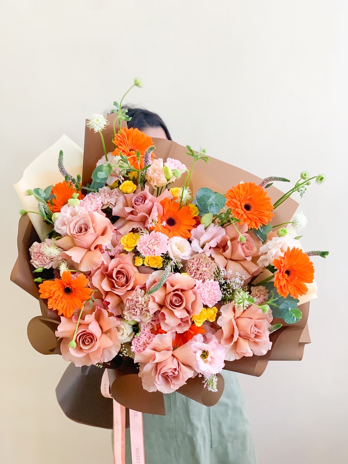 Large bouquet of peach roses, orange gerberas, pink carnations, and yellow mini roses wrapped in brown paper.