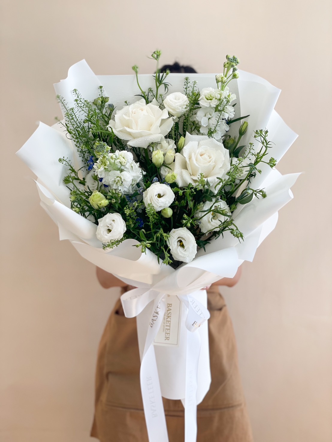 A luxurious bouquet featuring white roses, lisianthus, and greenery elegantly wrapped in white paper and tied with a Basketeer signature ribbon.
