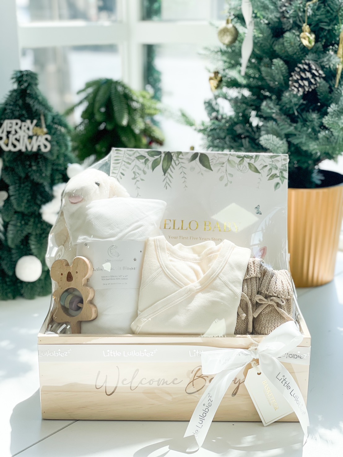 Wooden baby gift box with a muslin blanket, baby onesie, knitted booties, a wooden teether, and a "Hello Baby" memory book, tied with a white ribbon.