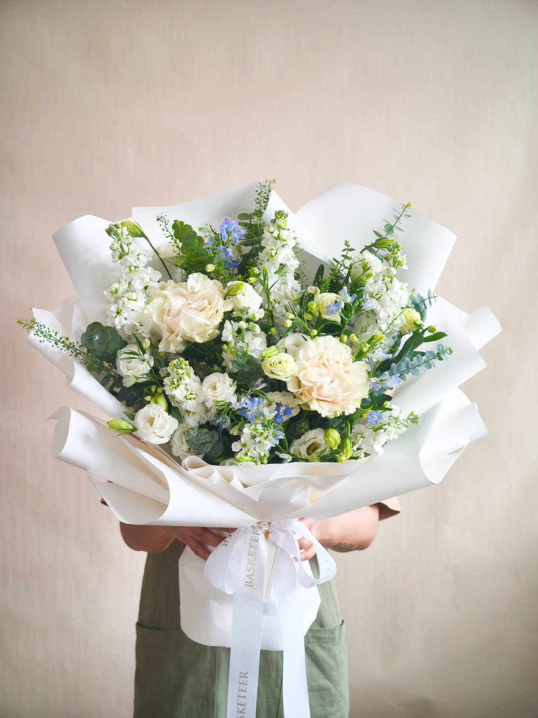 Elegant bouquet with cream roses, white lisianthus, blue delphiniums, hydrangeas, and greenery wrapped in white paper.