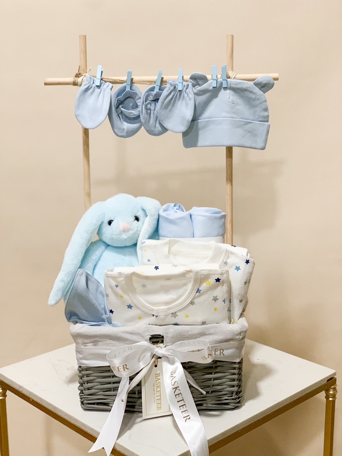 A wicker basket filled with Baby's First Comfort items against a light blue background. The contents include a blue stuffed bunny, blue blanket, baby onesie, blue socks, pacifiers, and wooden toys, all neatly arranged to create a charming and cozy presentation.
