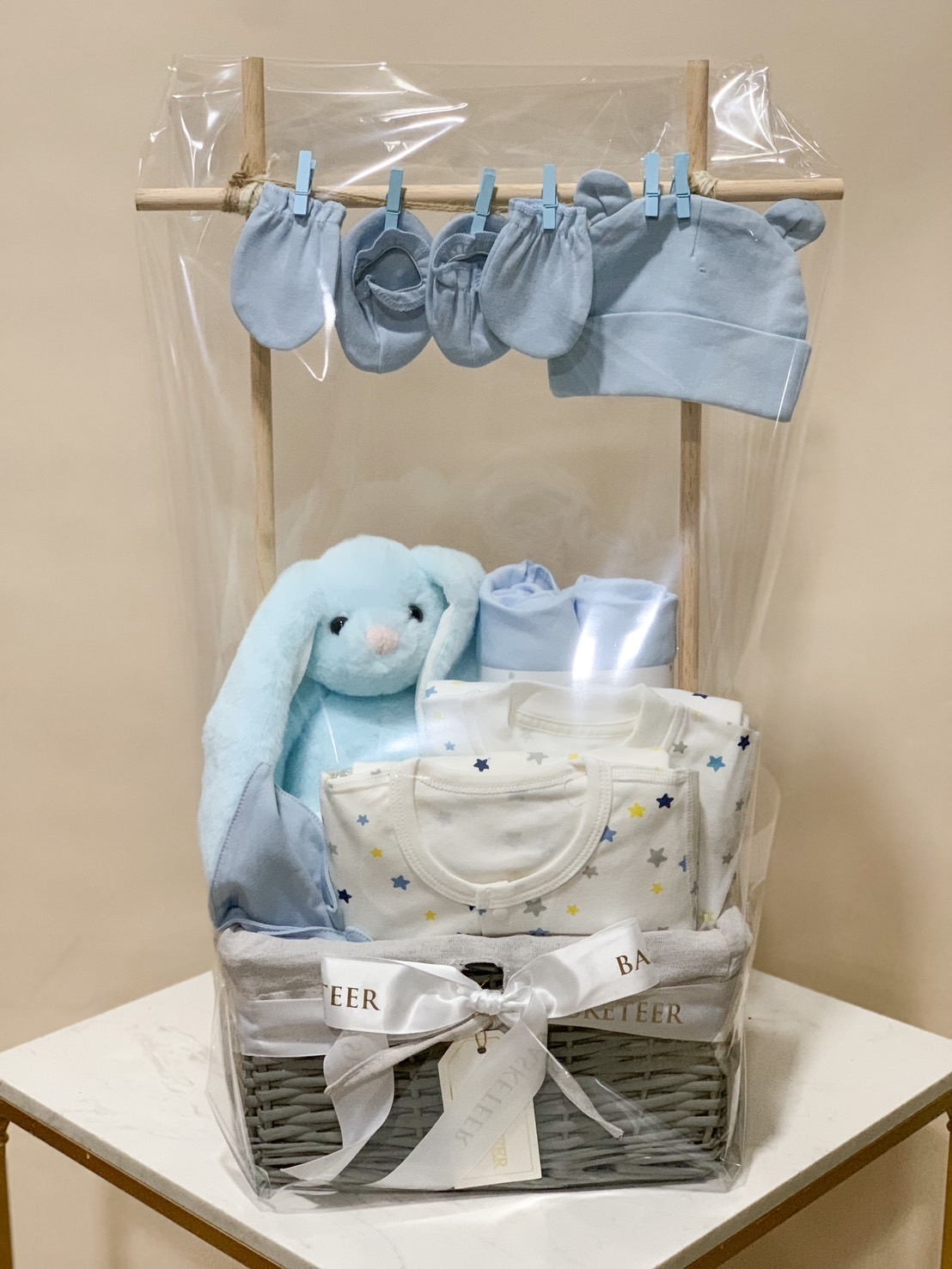 A wicker basket filled with Baby's First Comfort items against a light blue background. The contents include a blue stuffed bunny, blue blanket, baby onesie, blue socks, pacifiers, and wooden toys, all neatly arranged to create a charming and cozy presentation.