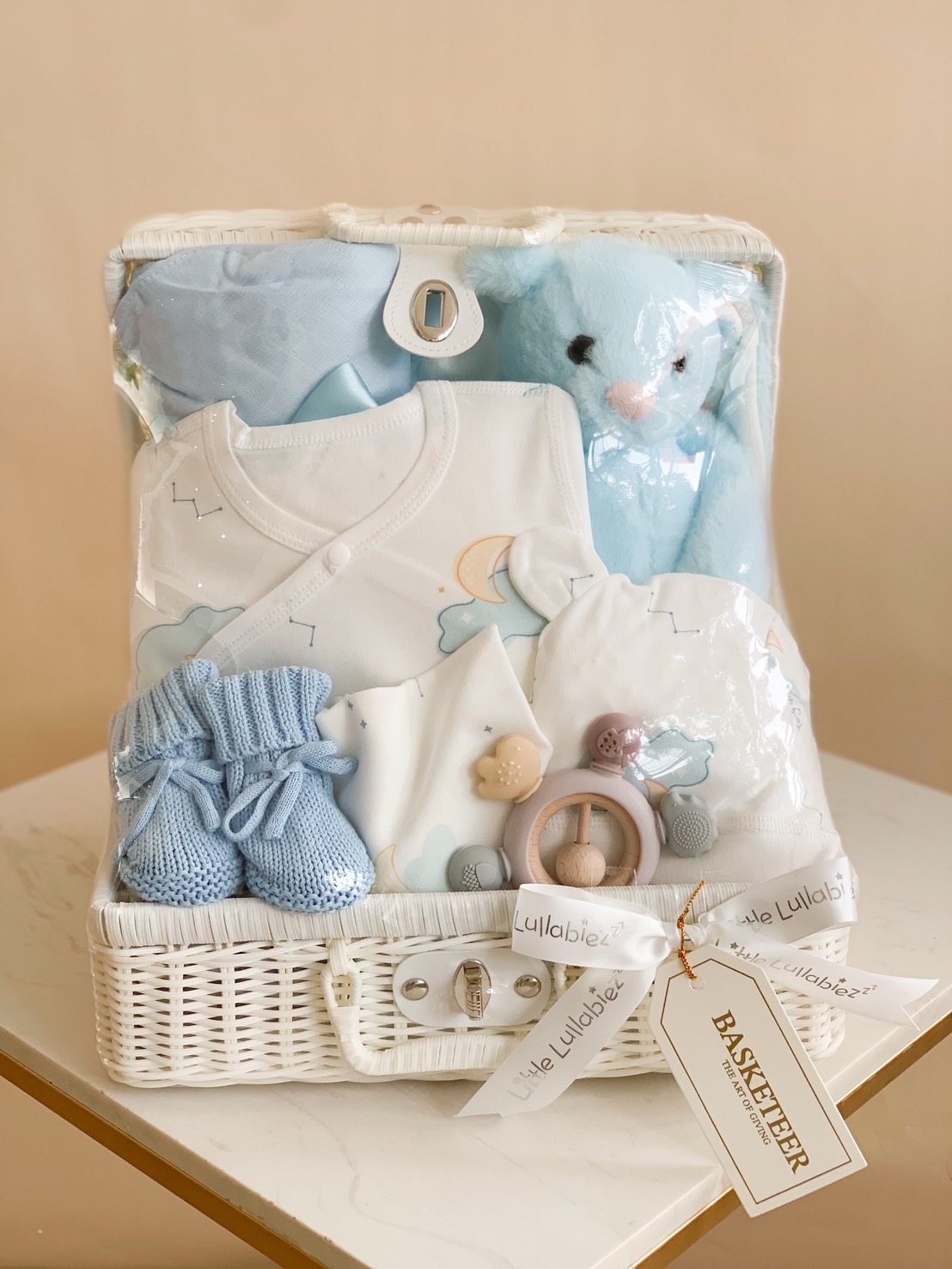 A gift basket with Baby's First Essentials includes a stuffed blue bunny, baby hats, mittens, booties, and onesies decorated with stars. The items are predominantly blue and white, and a ribbon around the basket is labeled 