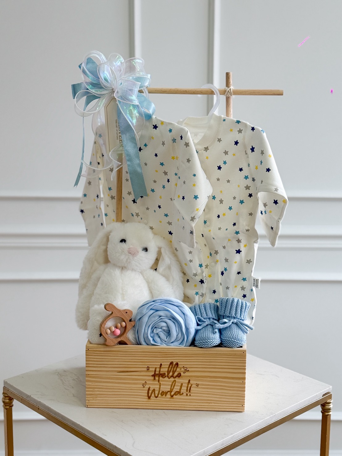 A baby gift set in a wooden box labeled “Hello World!!” featuring two star-patterned baby outfits hanging on a wooden frame, a white plush bunny, a blue baby blanket, blue knitted booties, and a wooden rattle. The decoration is completed with a blue and white ribbon bow on top.