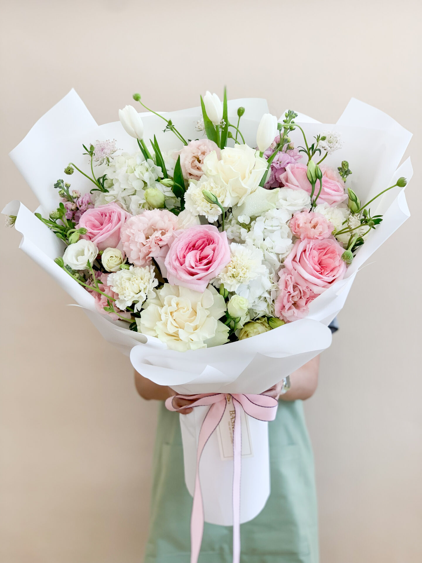 A person in a light brown apron holds a large bouquet of flowers wrapped in white paper. The bouquet features pink roses, pink gerbera daisies, white carnations, and greenery. A white ribbon with text is tied around the base of the bouquet.