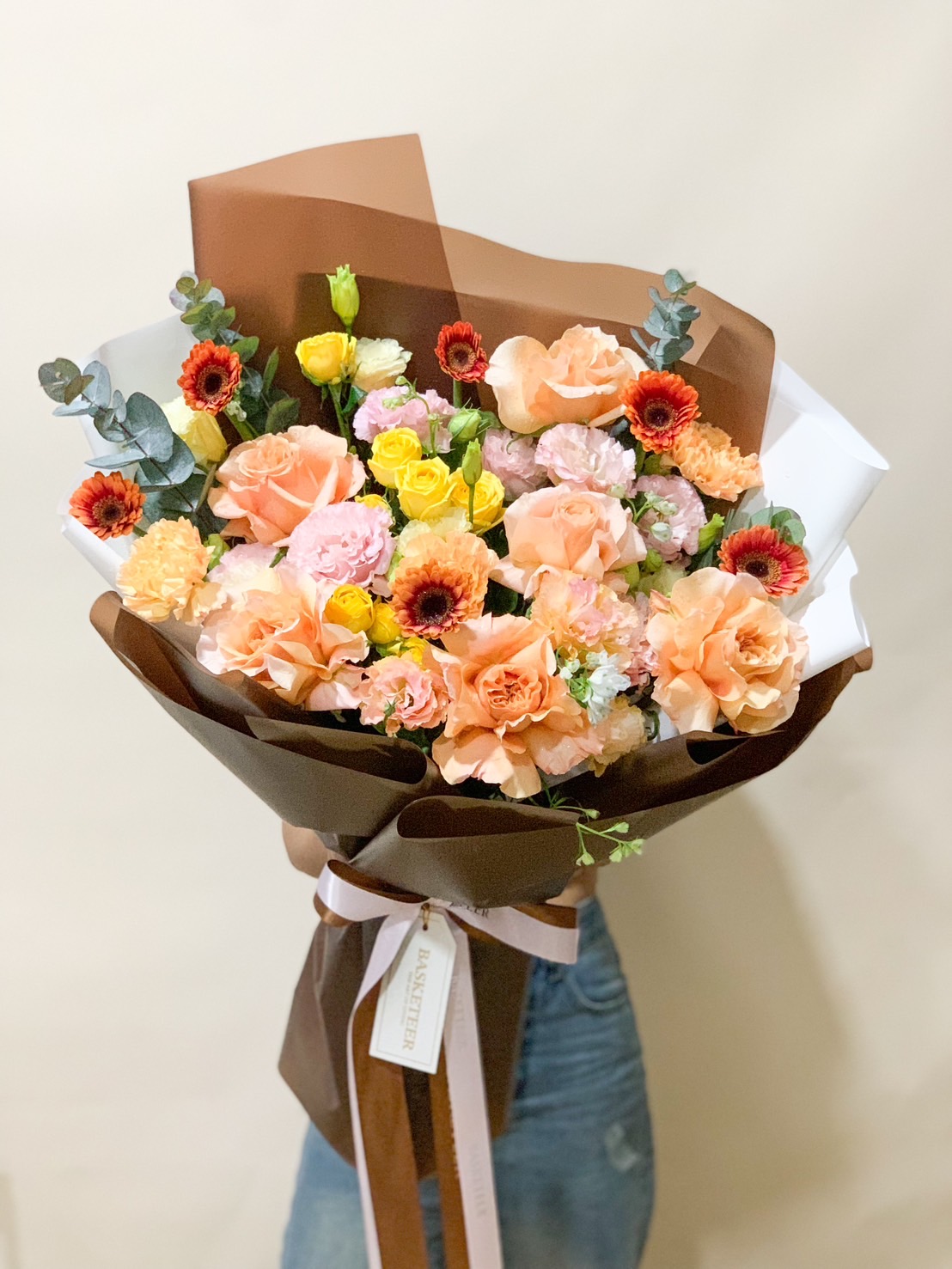 Elegant bouquet of peach roses, yellow blooms, and pink flowers, wrapped in brown and white paper with a luxury ribbon.
