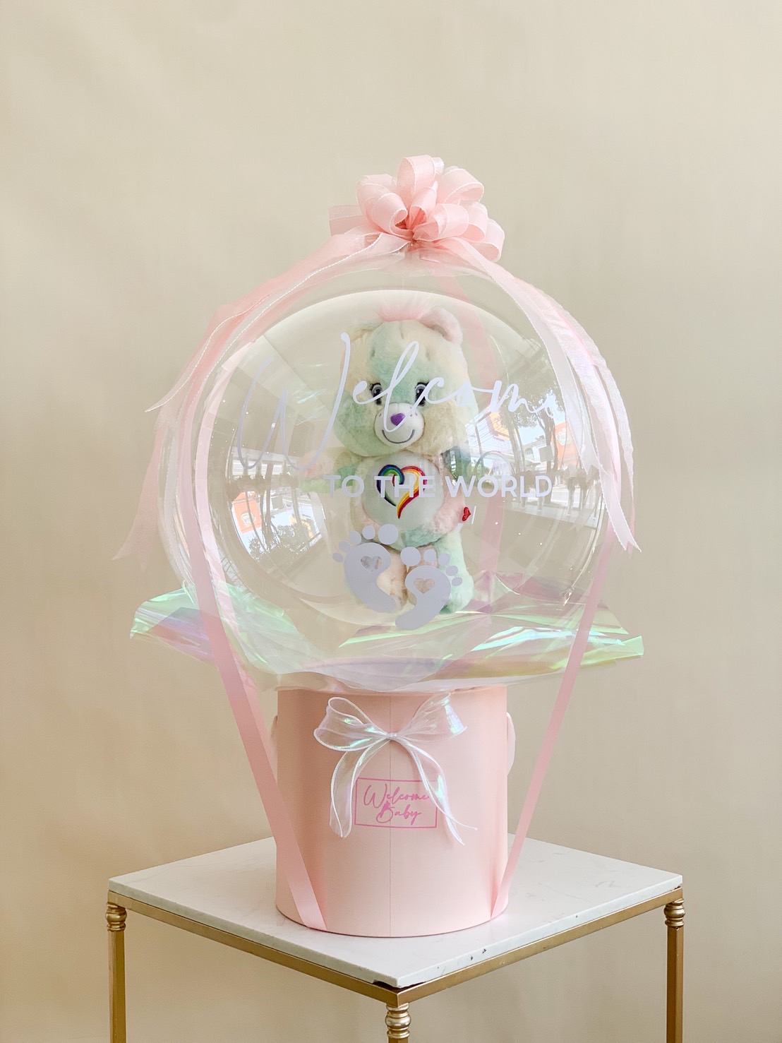 Clear welcome baby balloon with pastel teddy bear inside, pink ribbons, and a pink gift box for newborn celebration.