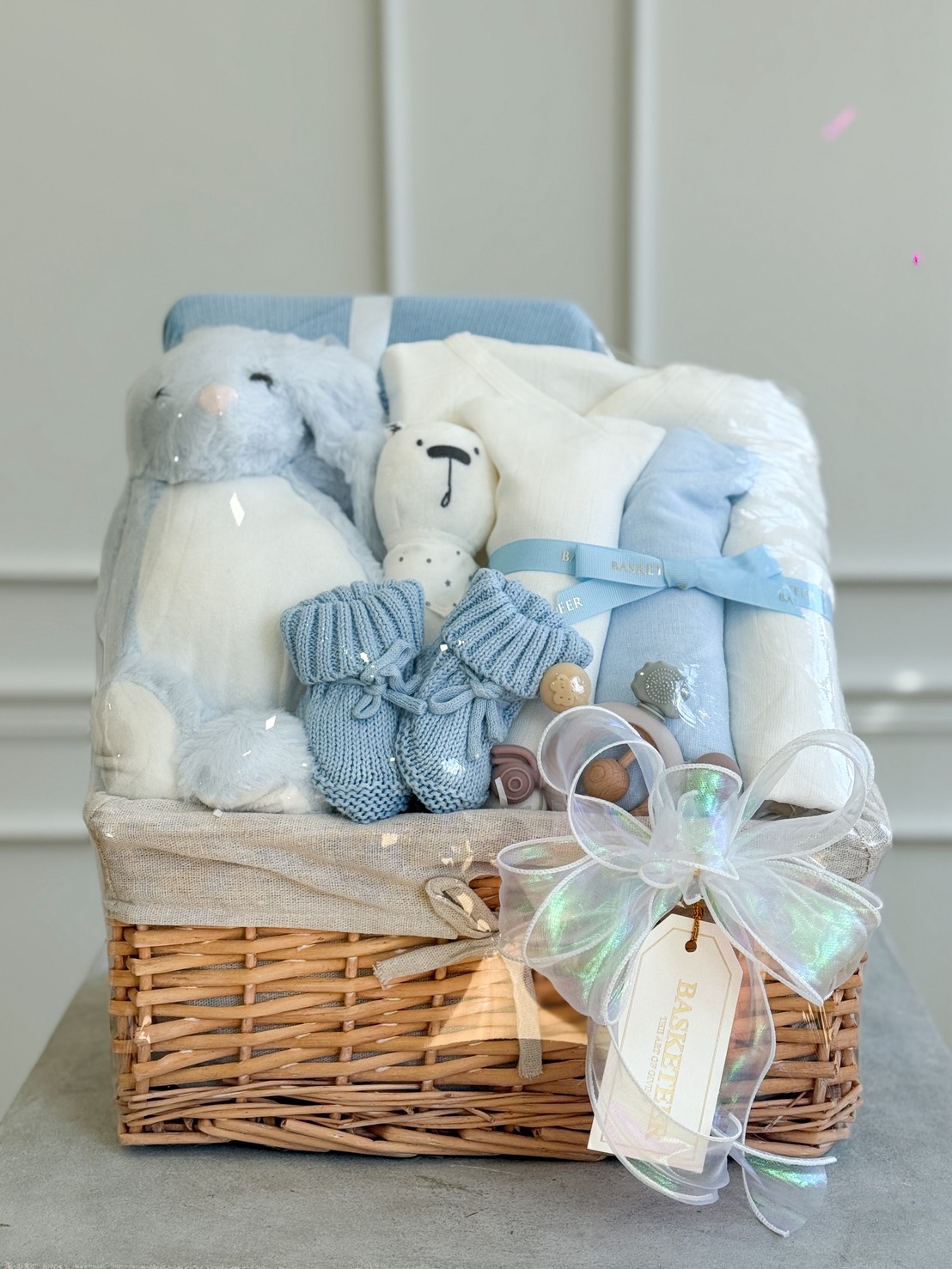 Luxury baby gift basket in blue tones featuring soft plush toys, baby clothes, knitted booties, and a white iridescent ribbon bow.