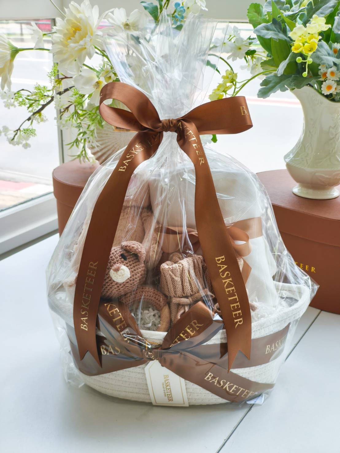 Baby gift basket wrapped in premium clear film with plush toy, knitted teddy, blanket, baby booties, and satin ribbon.