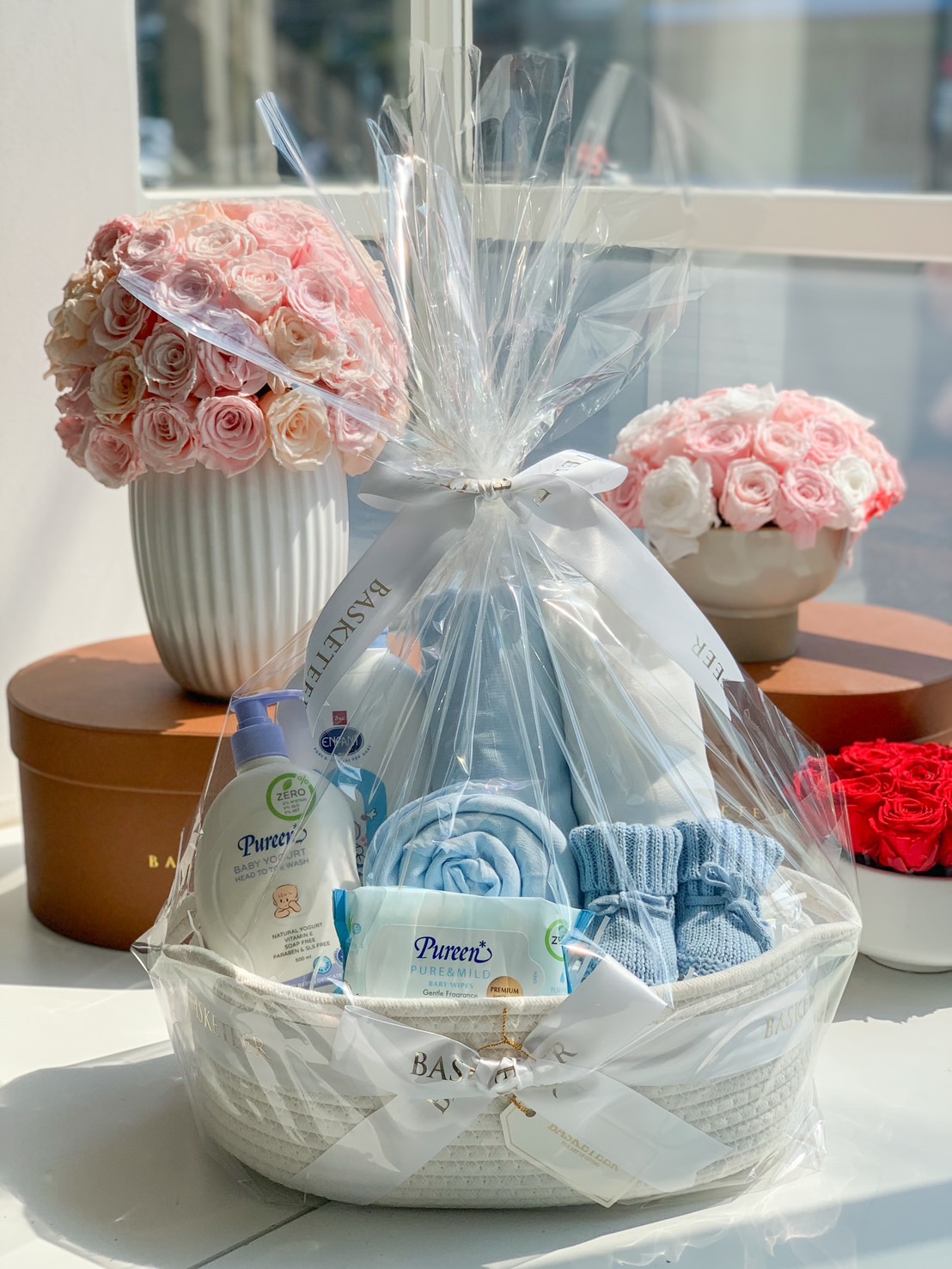 A beautifully wrapped blue-themed baby boy gift basket with Pureen baby care products, soft blankets, baby socks, and accessories in a stylish white woven basket.