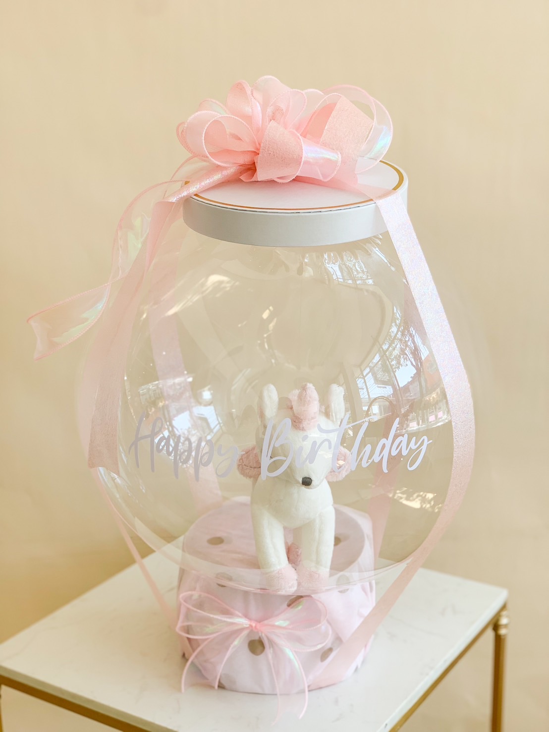 A beautifully curated birthday gift, featuring a clear bubble balloon with a plush toy inside, adorned with delicate pink ribbons and elegant details. A unique and heartwarming surprise for any special occasion.
