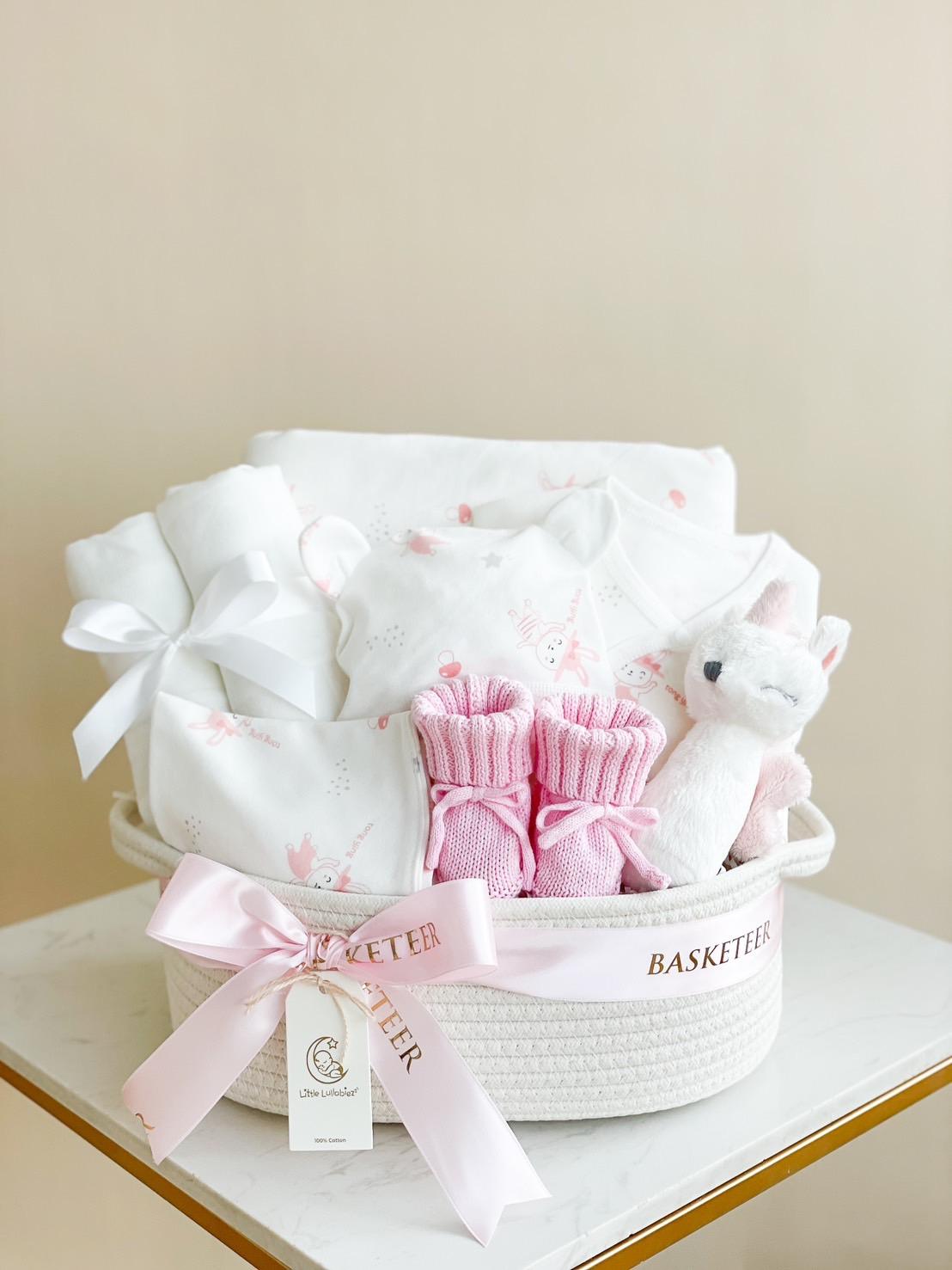A gift basket labeled "BASKETEER," this Pink Baby Girl Gift Basket is filled with baby essentials. It contains a plush toy, pink baby socks, white onesies with pink designs, and items wrapped with white ribbon. The basket is placed on a white surface against a neutral background.