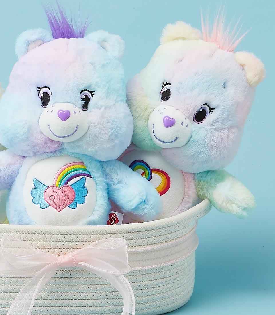 Two Care Bear plush toys sitting in a woven basket, one rainbow-colored and the other blue.