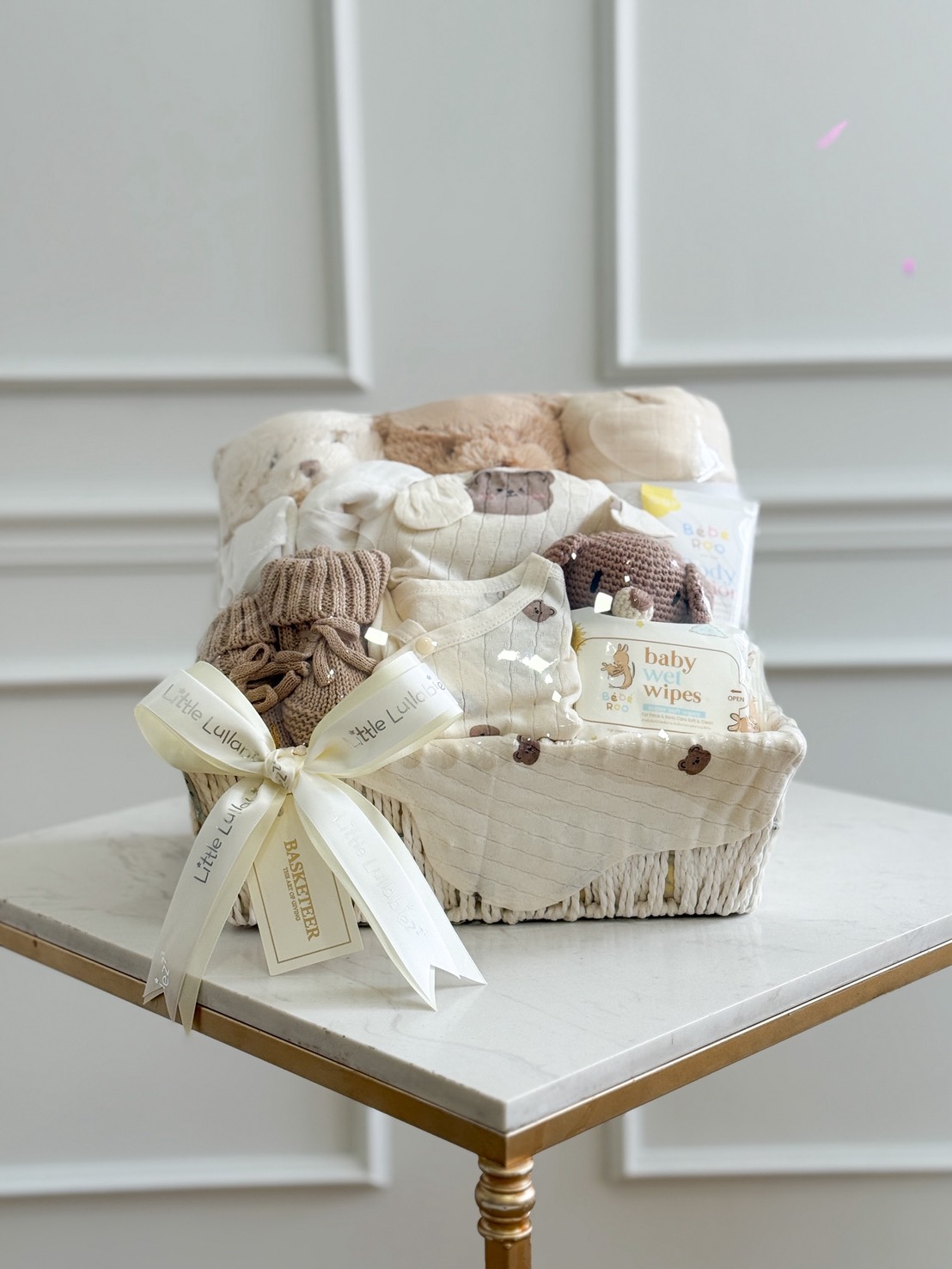 A white Bunny Friendly Baby gift hamper on a table holds neatly folded baby clothes, knitted hats, baby wipes, and a soft toy. The hamper is tied with white ribbon and displayed against a white-panelled wall.