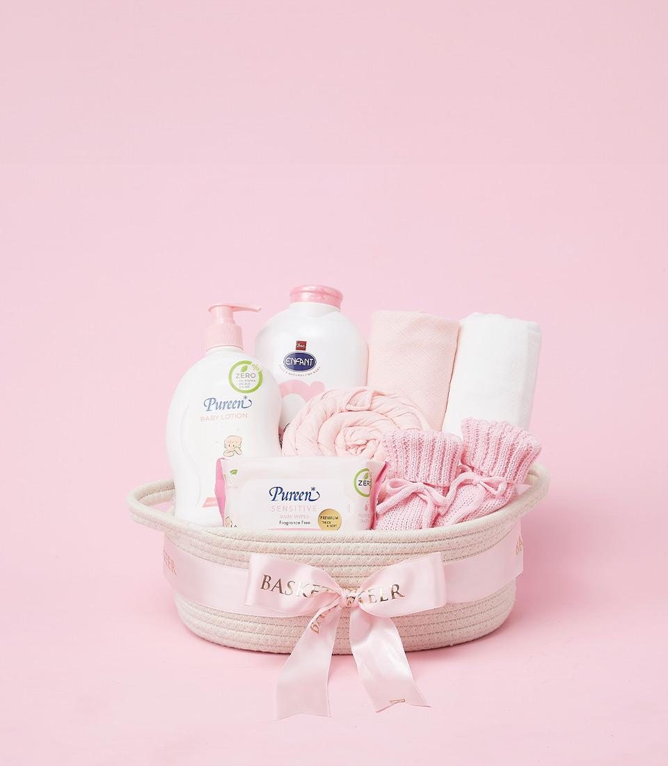 The Pink Pamper Basket, set against a matching pink background, is filled with essential baby care items. Inside, you'll find baby lotion, baby powder, a bar of baby soap, a folded white towel, a pink loofah, and pink knitted apparel. The basket is beautifully adorned with a ribbon.
