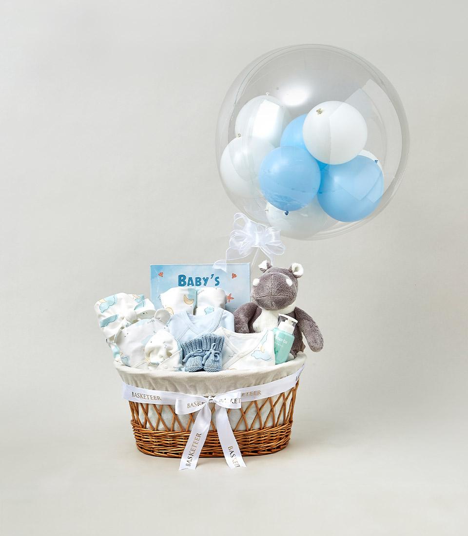 A baby gift basket with a clear balloon filled with blue and white balloons, featuring essential items like a hippopotamus plush toy, baby clothes