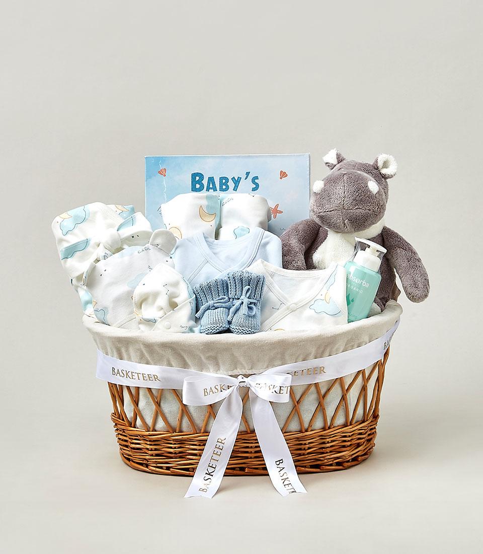 A wicker basket filled with baby gifts, including a gray stuffed bear, baby clothes, a baby book titled 