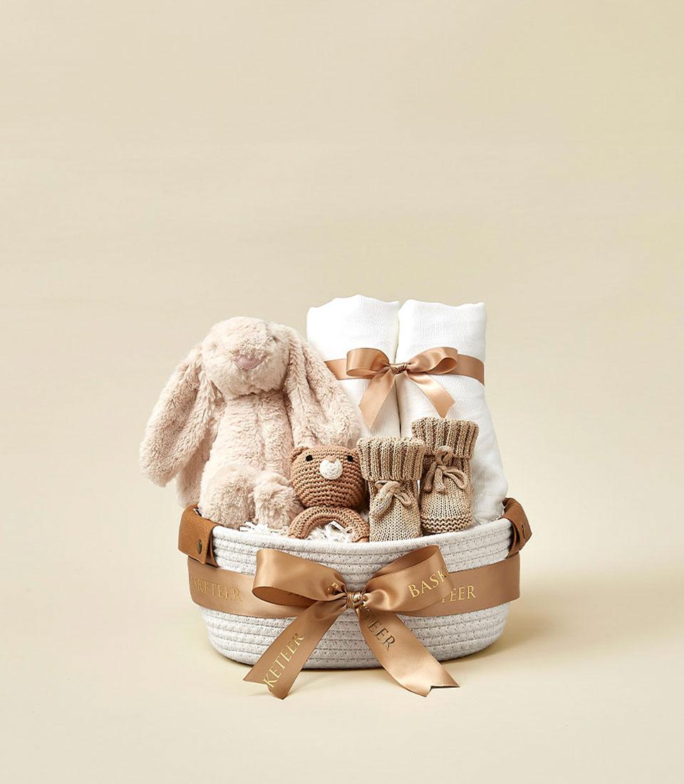 A neatly wrapped gift basket, a bundle of joy for your little one, contains a plush bunny toy, baby socks, a soft brown bear toy, white rolled-up baby blankets, and a beige and white woven basket. The basket is adorned with a brown ribbon that reads 