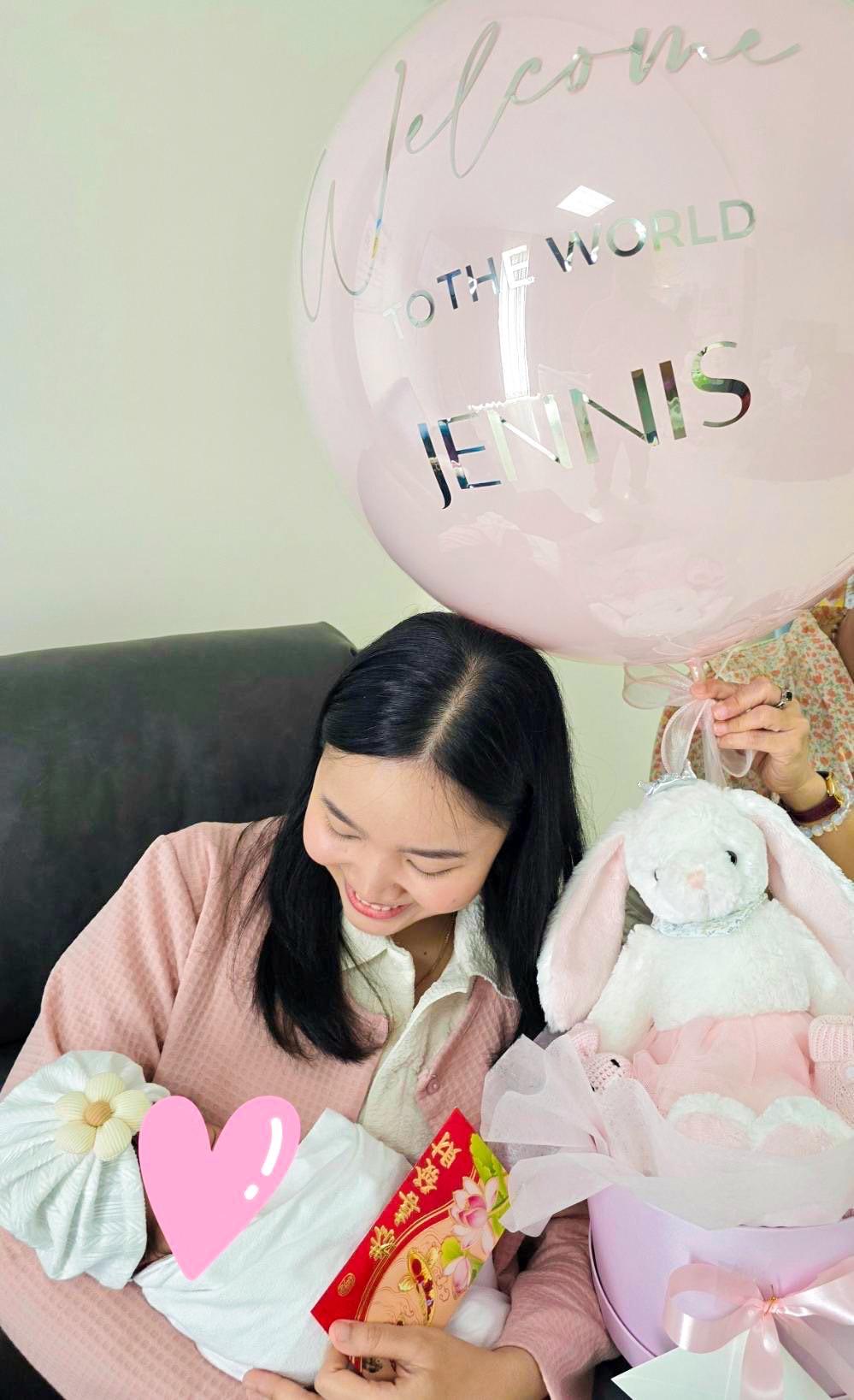 A woman holds a newborn baby wrapped in a white blanket with a heart emoji covering the baby's face. A large pink balloon with 
