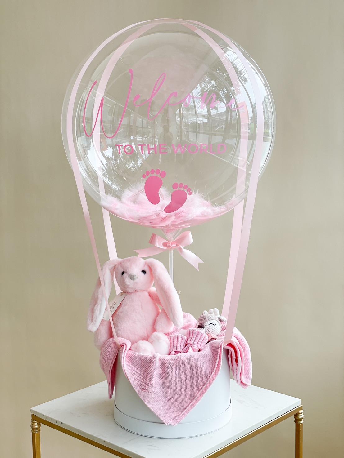 A newborn luxury gift, this decorative arrangement features a transparent balloon with "Welcome to the World" and pink baby footprints, attached by pink ribbons to a white base. Inside the base are a pink plush bunny and pink baby clothes, all atop a small white table.