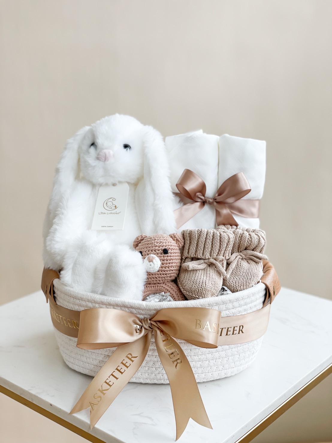 A white gift basket with the label "Basketeer" offers a bundle of joy for your little one, filled with baby items including a plush white rabbit, white towels tied with satin ribbons, knitted baby booties, a small knitted bear, and a pair of fuzzy slippers. The basket rests on a white surface.