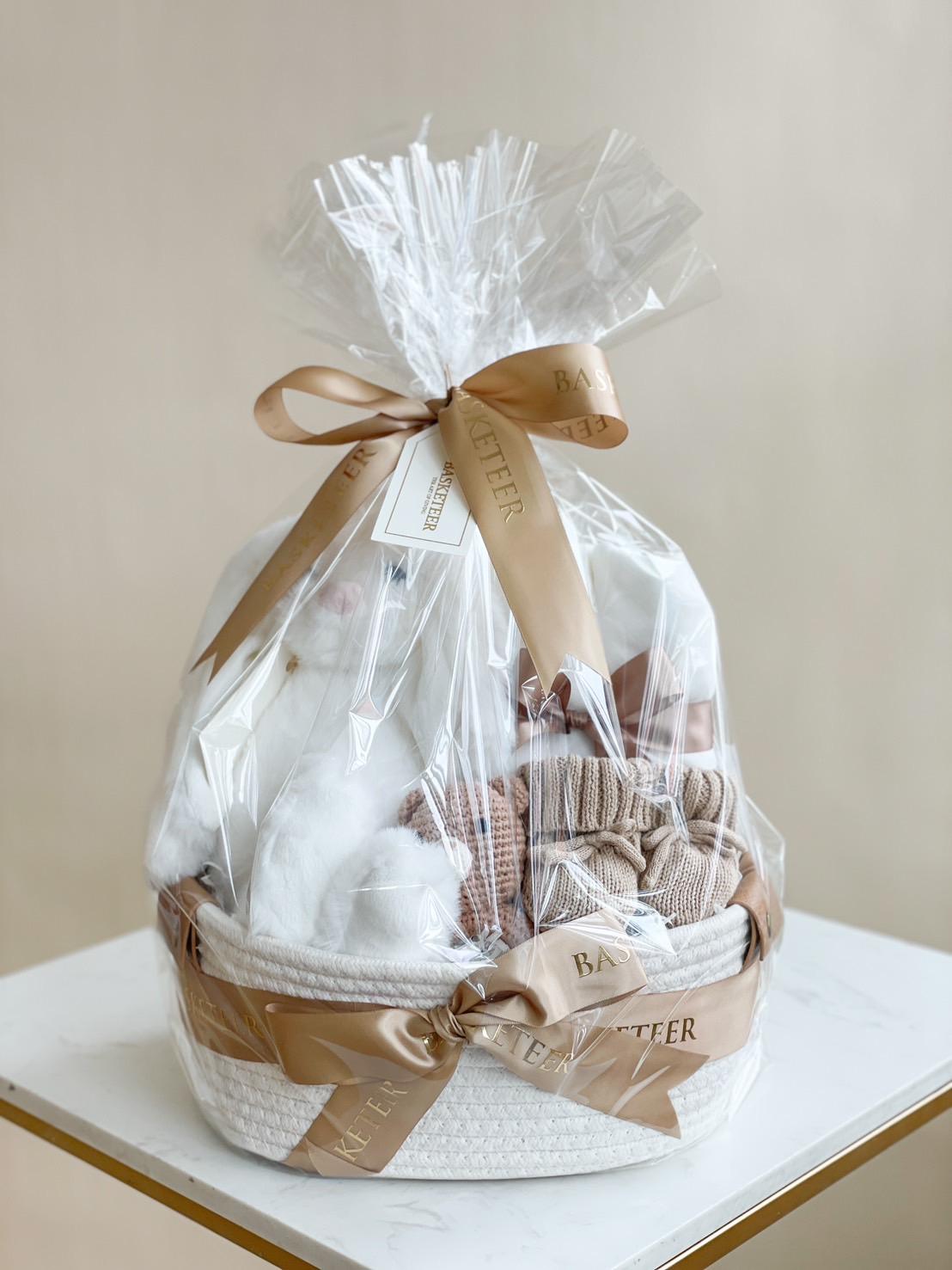 A beautifully wrapped gift basket, a bundle of joy for your little one, sits on a white surface. Enclosed in clear cellophane and tied with a beige ribbon, the basket contains various items in neutral tones, including plush toys and fabric goods. A small tag is attached to the ribbon.
