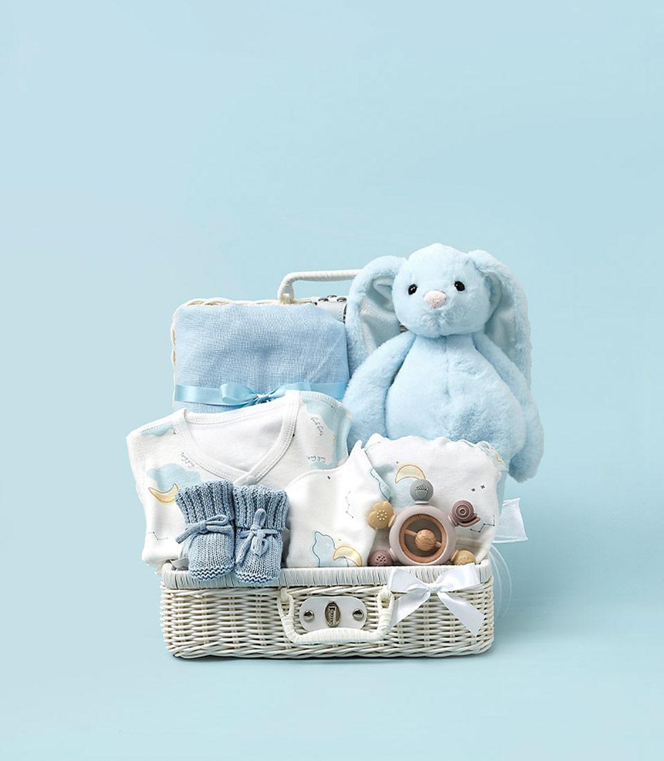 A wicker basket filled with Baby's First Comfort items against a light blue background. The contents include a blue stuffed bunny, blue blanket, baby onesie, blue socks, pacifiers, and wooden toys, all neatly arranged to create a charming and cozy presentation.