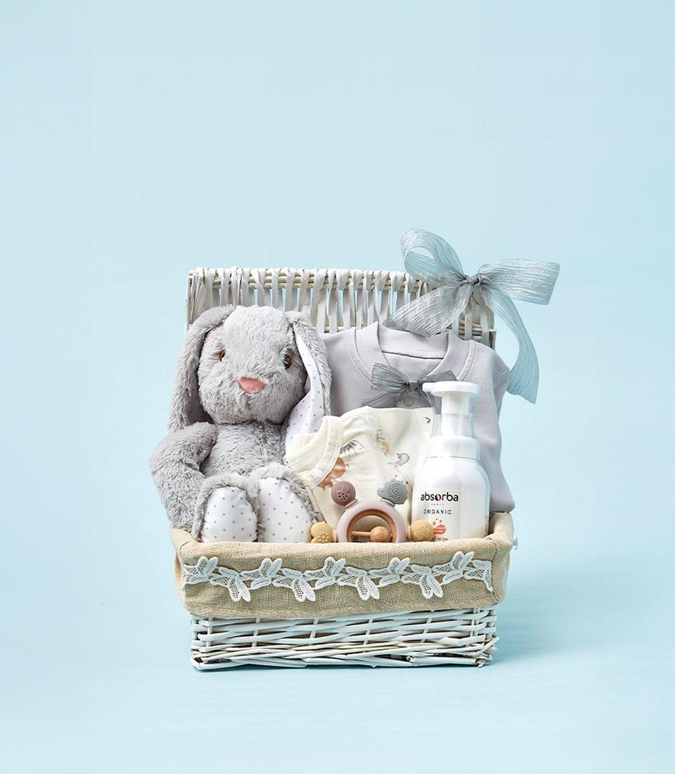 A white wicker basket with a grey ribbon bow, A Gift from the Heart, contains a stuffed grey bunny, a baby onesie, a pacifier, a wooden rattle, and a bottle of baby lotion. The background is light blue.