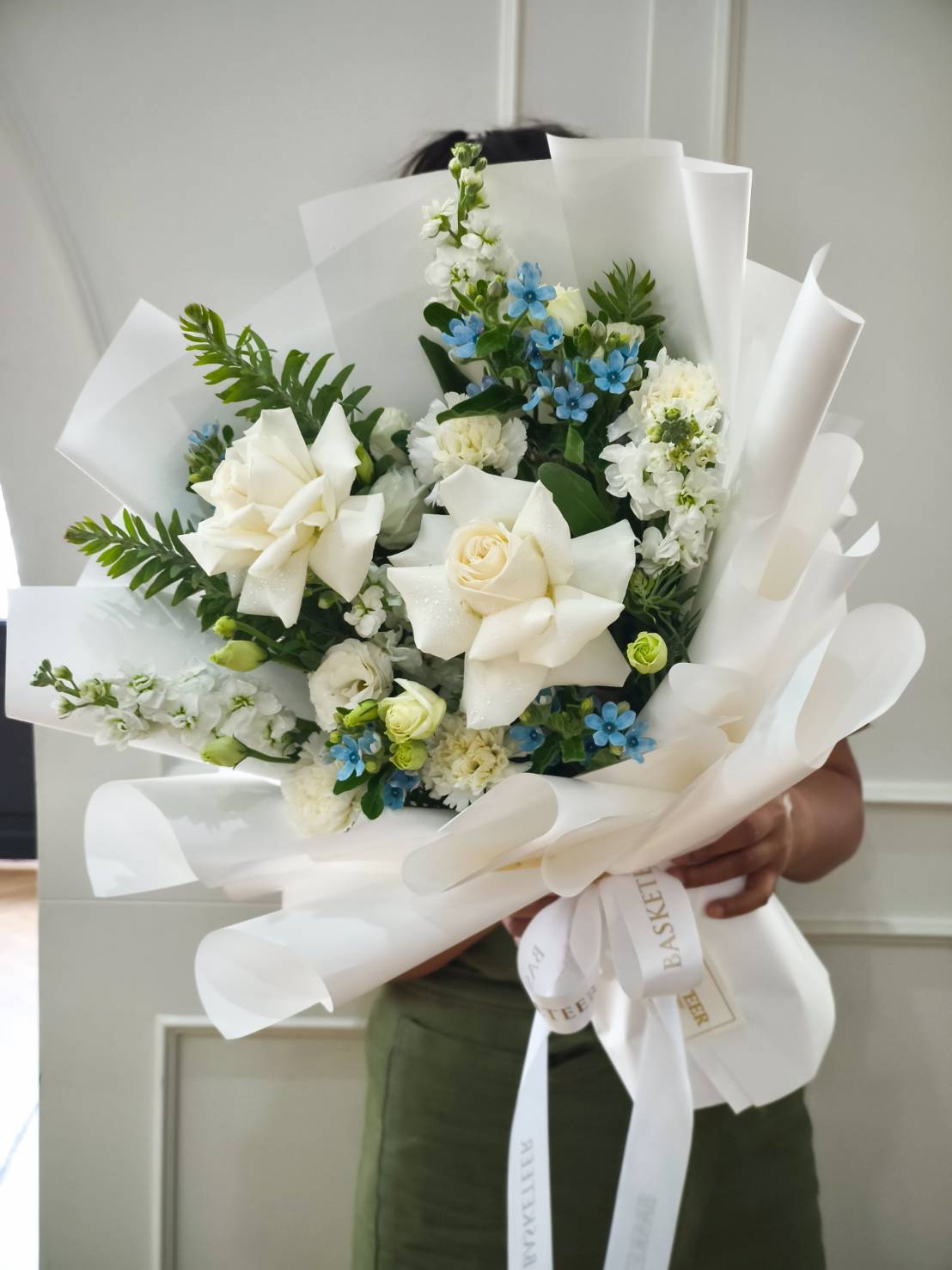 Elegant bouquet with white roses, carnations, and blue accent flowers, wrapped in crisp white paper with satin ribbon