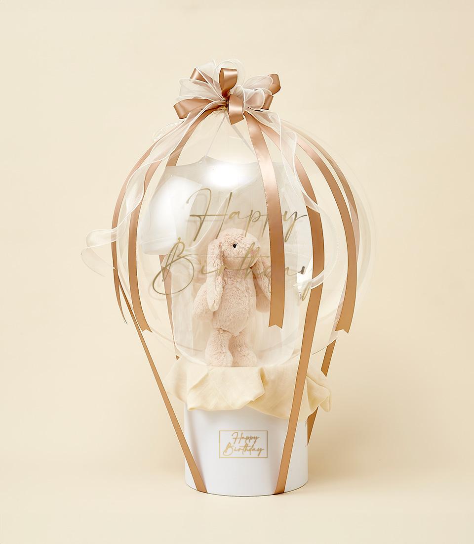 A transparent hot air balloon-shaped gift with a beige plush bunny inside. The balloon is tied with elegant white and gold ribbons, and stands in a white box labeled 