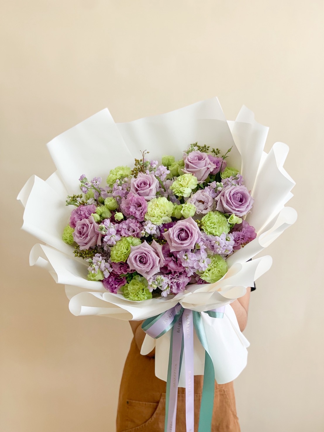 Bouquet of lavender roses, light green carnations, and lilac fillers wrapped in white paper with pastel ribbons.