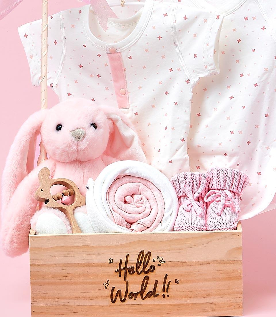 A pink wooden gift basket filled with baby clothes, a plush bunny toy, a rattle, and a swaddle blanket.