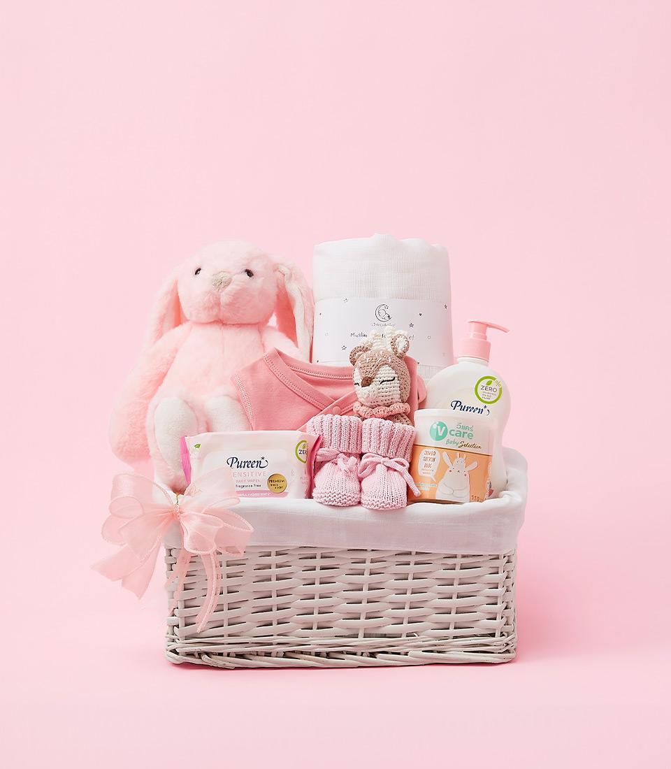 A pink wicker basket filled with baby essentials, including a plush bunny, clothes, a towel, and baby care products.