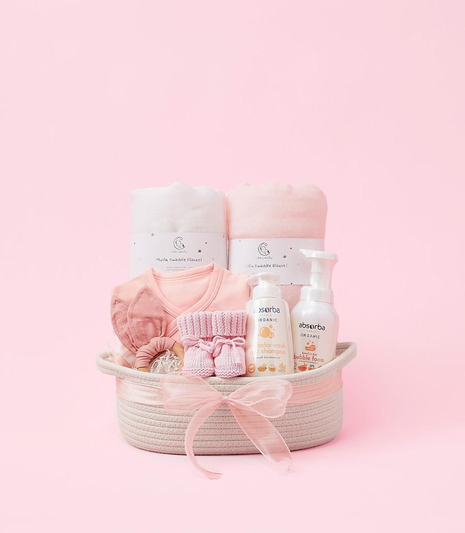 A pink woven basket filled with baby essentials, including swaddle blankets, clothes, a rattle, and baby care products.
