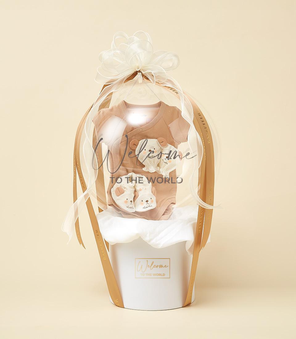 A white gift basket resembling a hot air balloon, adorned with ribbons and a bow, serves as the perfect gift for a new life. Inside, it contains baby items including a beige onesie and small shoes. The basket's ribbon features the words 