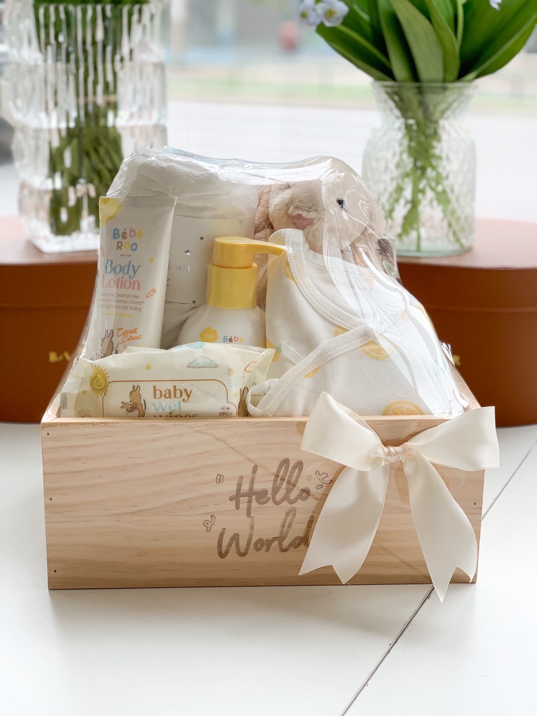 Newborn baby gift box with lotion, baby clothes, wipes, and plush toy in a wooden “Hello World” crate.