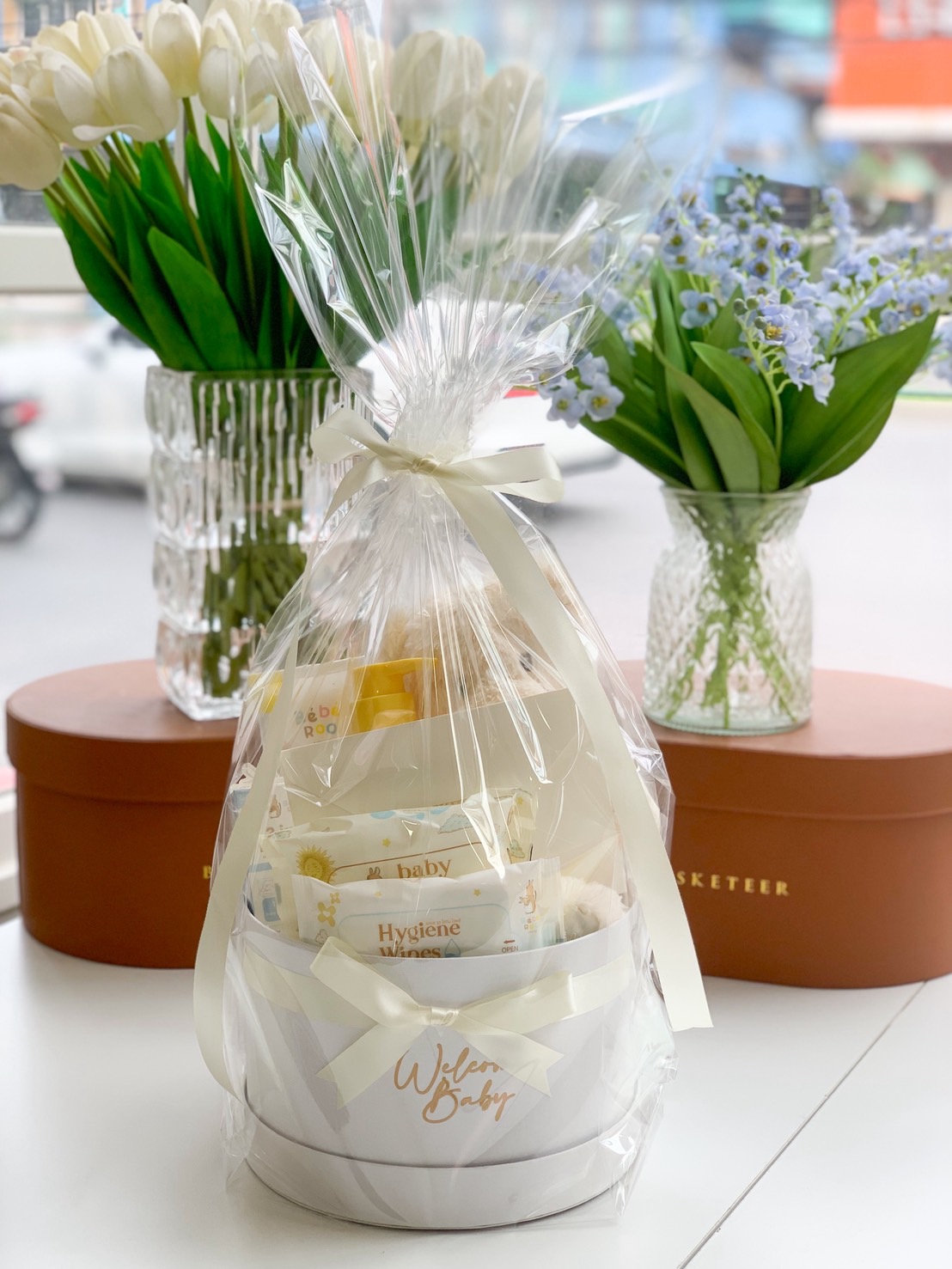 White round baby gift box wrapped in clear plastic, filled with baby wipes, lotion, wash, and plush toy.