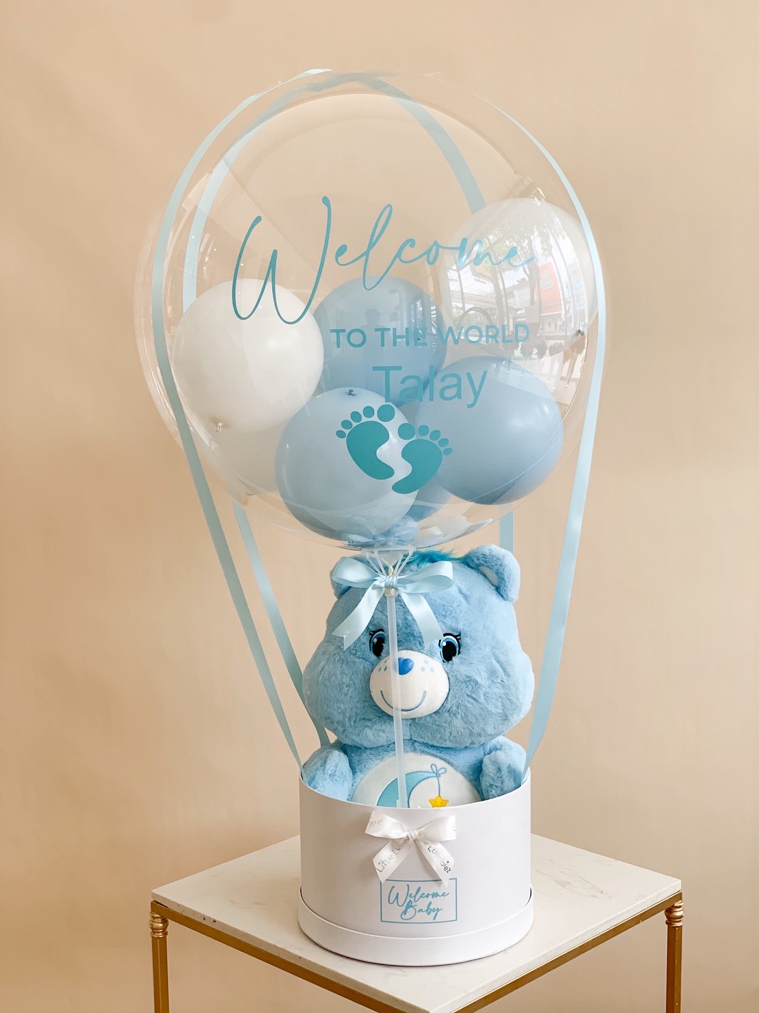 Baby blue teddy bear in a round white gift box topped with a clear balloon reading “Welcome to the World Talay.”
