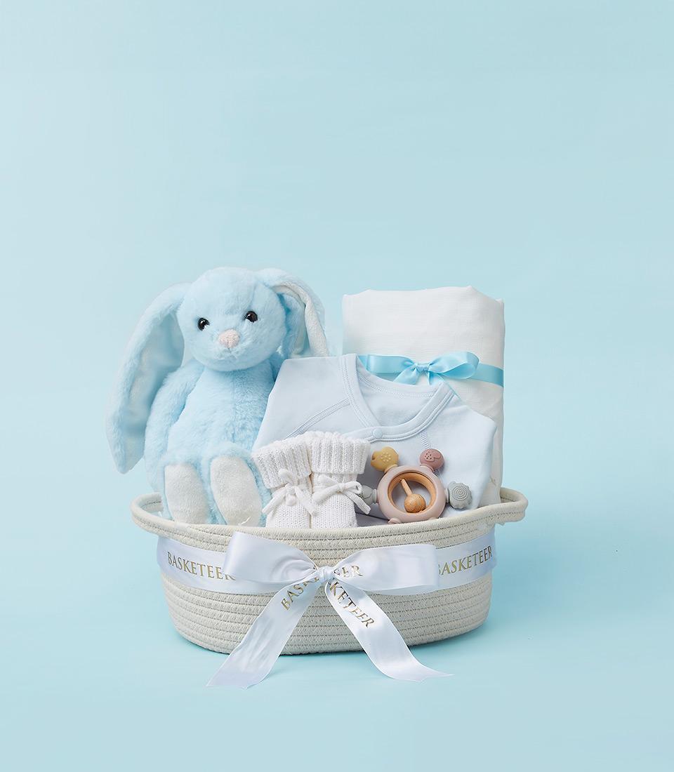 A blue woven basket filled with baby essentials, including a plush bunny, clothes, a blanket, and baby care products.