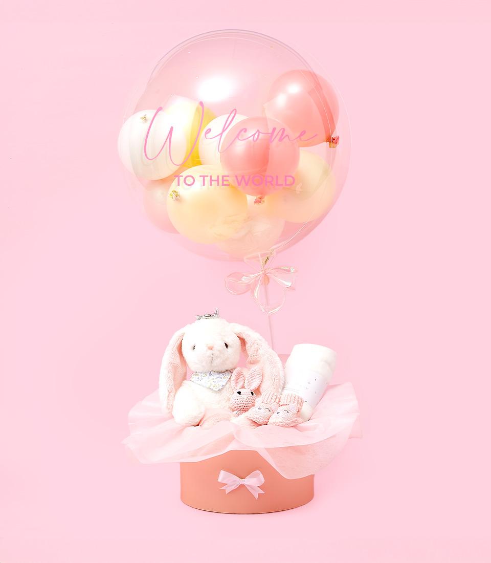 A pastel pink gift box contains baby essentials like pink booties, a white onesie, and a stuffed bunny. Above it, a transparent balloon filled with pink and yellow mini balloons reads 